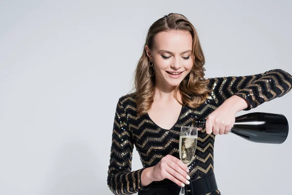 Heureuse jeune femme tenant verre tout en versant du champagne de bouteille isolé sur gris — Photo de stock