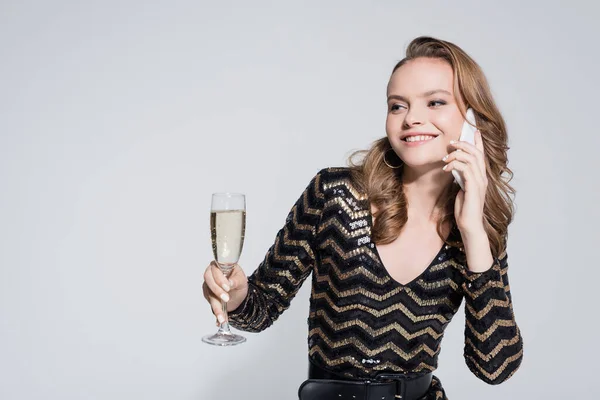 Lächelnde junge Frau hält ein Glas Champagner in der Hand und spricht auf dem Smartphone isoliert auf grau — Stockfoto