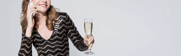 Vista recortada de una mujer sonriente sosteniendo una copa de champán y hablando en un teléfono inteligente aislado en gris, pancarta - foto de stock