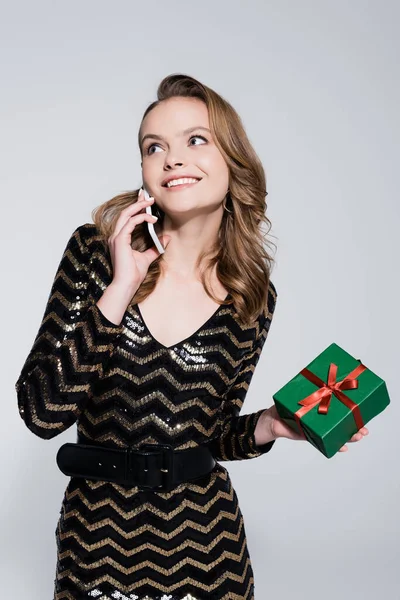 Lächelnde junge Frau hält Weihnachtsgeschenk in der Hand und telefoniert auf Smartphone isoliert auf grau — Stockfoto
