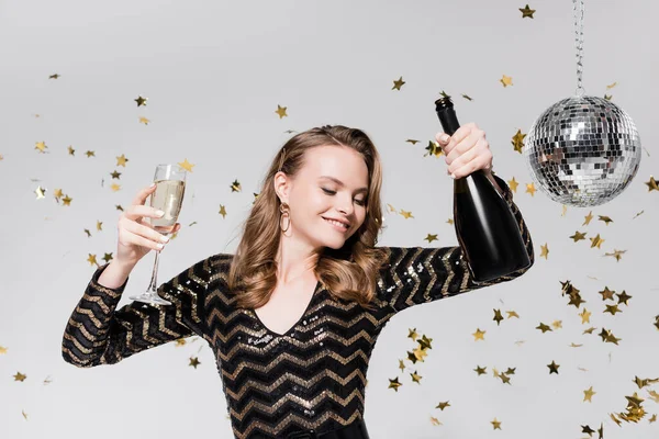 Alegre joven mujer sosteniendo copa y botella de champán cerca de la bola disco y la caída de confeti en gris - foto de stock