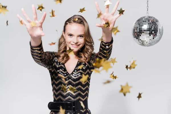 Happy young woman standing near disco ball and throwing confetti on grey — Stock Photo