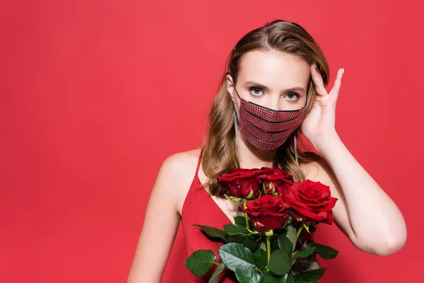 Молода жінка в захисній масці зі стразами тримає троянди і дивиться на камеру на червоному — стокове фото