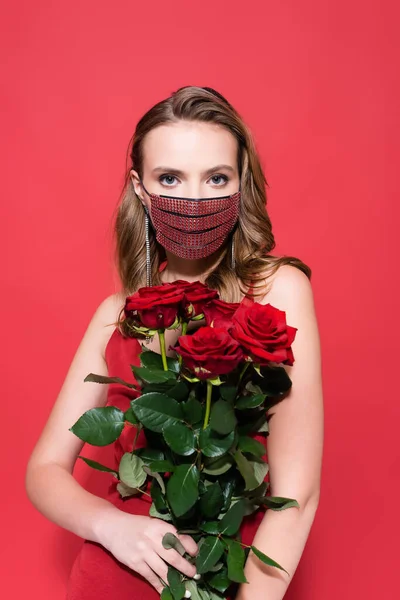 Mulher em máscara protetora com strass segurando rosas e olhando para a câmera no vermelho — Fotografia de Stock