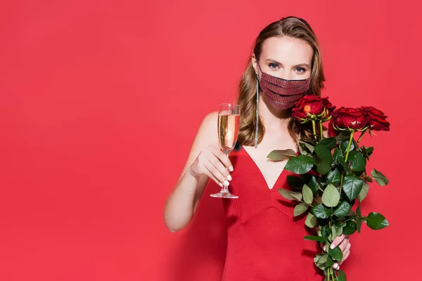Jovem mulher em máscara protetora com strass segurando copo de champanhe e rosas no vermelho — Fotografia de Stock