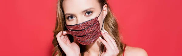 Young woman wearing protective mask with rhinestones and looking at camera on red, banner — Stock Photo