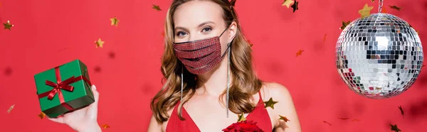 Woman in protective mask with rhinestones holding christmas present near confetti and disco ball on red, banner — Stock Photo