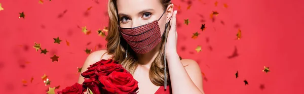 Woman in protective mask with rhinestones fixing hair near roses and confetti on red, banner — Stock Photo