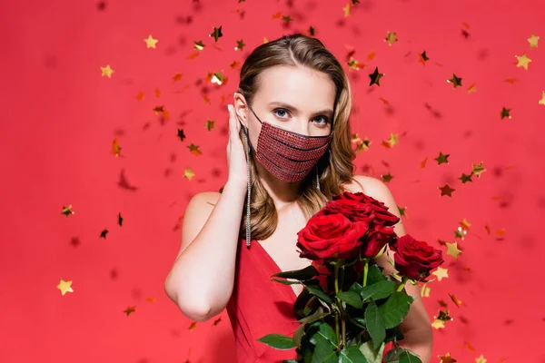 Femme en masque de protection avec strass fixant les cheveux près des roses et des confettis sur rouge — Photo de stock