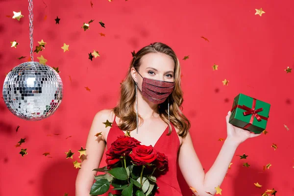 Femme en masque de protection avec strass tenant Noël présent près de confettis et boule disco sur rouge — Photo de stock