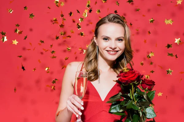 Glückliche Frau hält Rosen und ein Glas Champagner in der Nähe fallender Konfetti auf Rot — Stockfoto