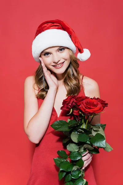 Happy young woman in santa hat holding roses on red — Stock Photo