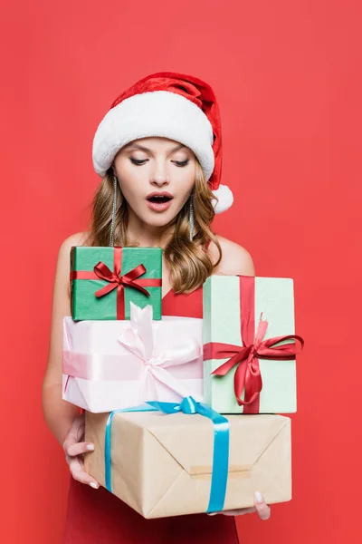 Femme surprise dans santa chapeau regardant des boîtes-cadeaux sur rouge — Photo de stock