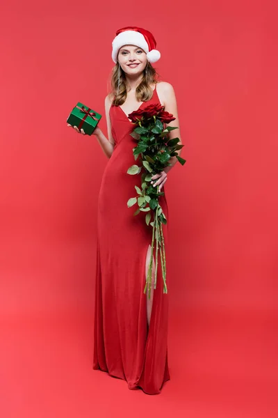 Longitud completa de la mujer feliz en vestido y sombrero de santa celebración de rosas y regalo de Navidad en rojo - foto de stock