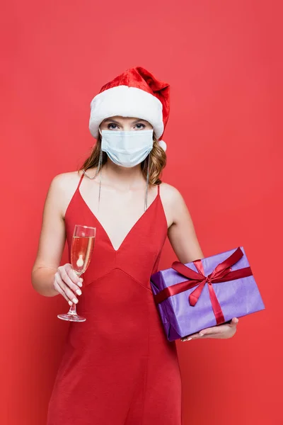 Jeune femme en santa chapeau et masque médical tenant cadeau de Noël et verre de champagne sur rouge — Photo de stock