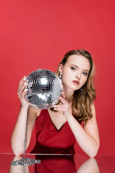 Joven mujer sosteniendo brillante bola disco y mirando a la cámara en rojo - foto de stock