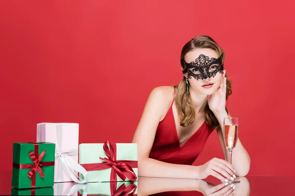Jeune femme en élégant masque de dentelle tenant verre de champagne près de cadeaux de Noël sur rouge — Photo de stock