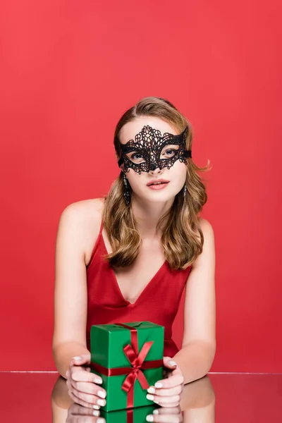 Jeune femme en élégant masque de dentelle tenant cadeau de Noël sur rouge — Photo de stock