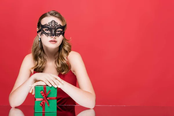 Young woman in black lace mask holding christmas present on red — Stock Photo