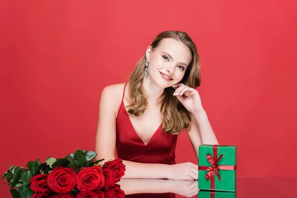 Молода щаслива жінка дивиться на камеру біля троянд і різдвяну подарункову коробку на червоному — стокове фото