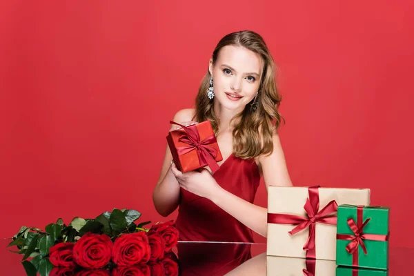 Smiling young woman holding christmas present near roses on red — Stock Photo