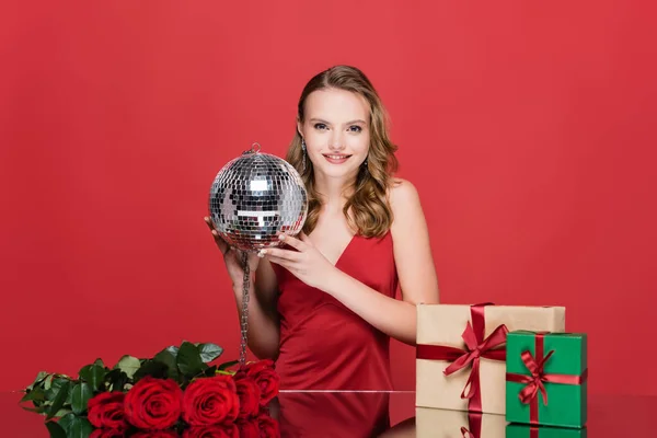 Mujer alegre sosteniendo bola disco cerca de regalos de Navidad y rosas en rojo — Stock Photo