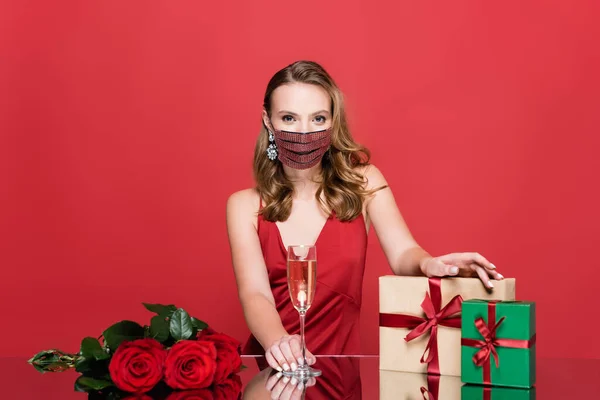 Frau in Schutzmaske mit Strass hält Glas Champagner in der Nähe von Rosen und Weihnachtsgeschenken auf Rot — Stockfoto