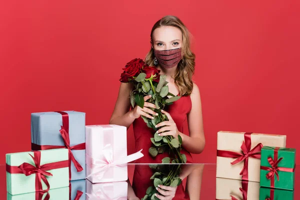 Femme en masque de protection avec strass tenant des roses près de cadeaux de Noël sur rouge — Photo de stock