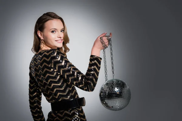 Happy young woman in dress looking at camera while holding disco ball on grey — Stock Photo
