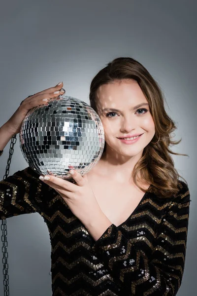 Cheerful young woman looking at camera while holding disco ball on grey — Stock Photo
