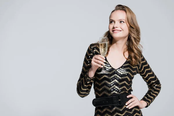 Cheerful woman holding glass of champagne and standing with hand on hip isolated on grey — Stock Photo