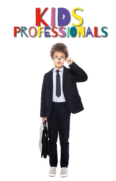 Chico sorprendido en traje fijación gafas y la celebración de maletín cerca de niños profesionales letras en blanco - foto de stock