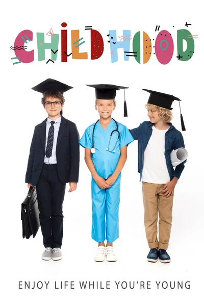 Children in graduation caps dressed in costumes of different professions standing near childhood lettering on white — Stock Photo