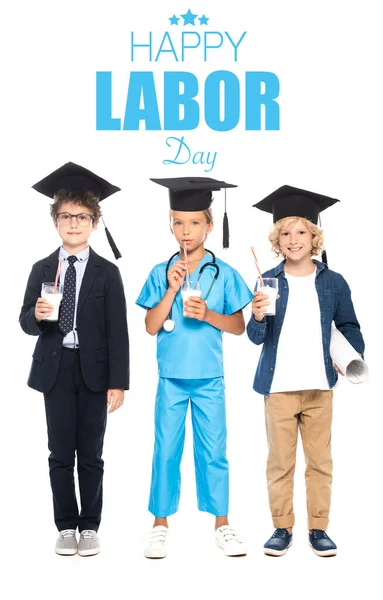 Niños en gorras de graduación vestidos con disfraces de diferentes profesiones sosteniendo vasos con leche cerca de letras felices del día del trabajo en blanco - foto de stock