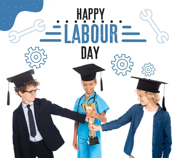 Kids in graduation caps dressed in costumes of different professions holding golden trophy near happy labour day lettering on white — Stock Photo
