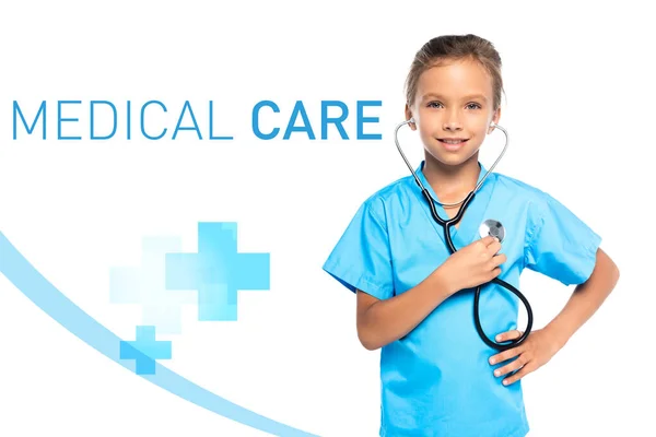 Child in costume of doctor holding stethoscope while standing with hand on hip near medical care lettering on white — Stock Photo