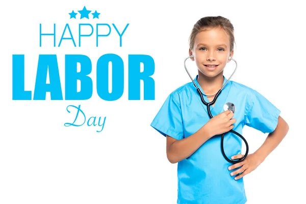 Child in costume of doctor holding stethoscope while standing with hand on hip near happy labor day lettering on white — Stock Photo