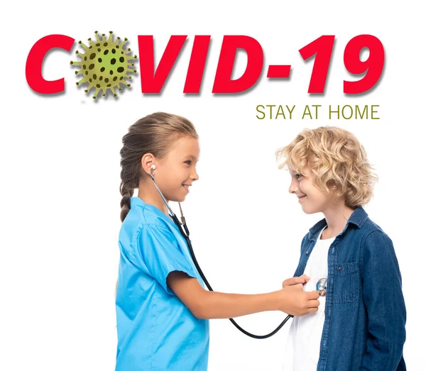 Side view of child in costume of doctor examining blonde boy near covid-19 stay at home lettering on white — Stock Photo