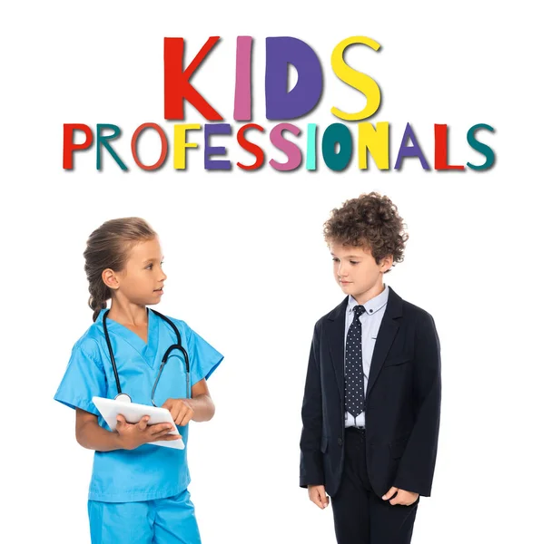 Child in costume of doctor using digital tablet near curly boy in formal wear and kids professionals lettering on white — Stock Photo