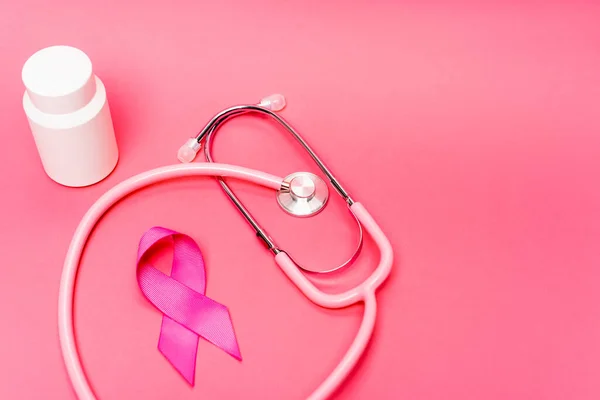 Stetoscopio vicino al nastro rosa di consapevolezza del cancro al seno e vaso con pillole su sfondo rosa — Foto stock