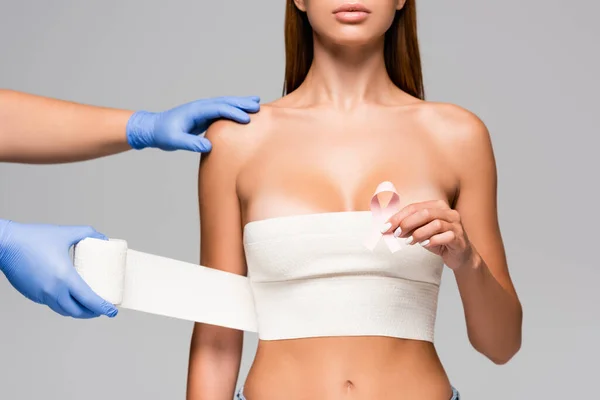 Vista recortada del médico envolviendo el pecho de una mujer joven con vendaje elástico mientras sostiene la cinta de la conciencia del cáncer de mama aislada en gris - foto de stock