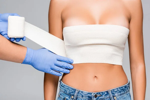 Vue recadrée du médecin en gants de latex enveloppant buste de jeune femme avec bandage élastique isolé sur gris — Photo de stock