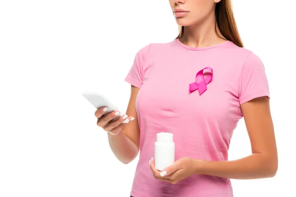 Vista ritagliata della donna con il nastro di consapevolezza del cancro al seno che tiene il vaso con le pillole e usando smartphone isolato su bianco — Foto stock