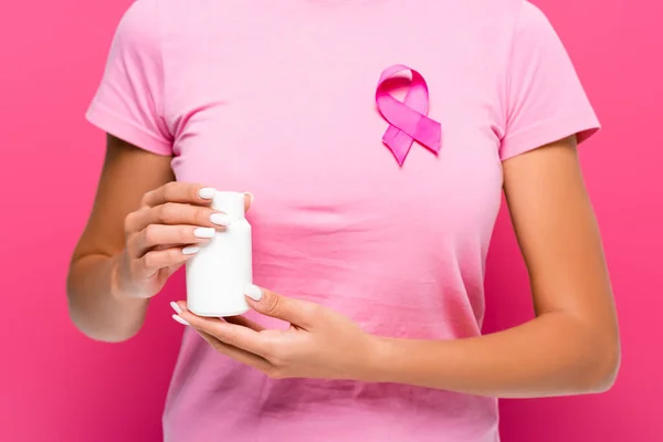 Vista recortada de una mujer joven sosteniendo frasco con pastillas mientras usa el signo de conciencia del cáncer de mama en el fondo rosa - foto de stock
