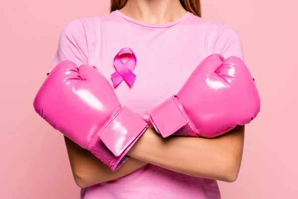 Partial view of woman in boxing gloves and ribbon of breast cancer awareness standing with crossed arms isolated on pink — Stock Photo