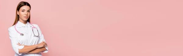 Молодий лікар з схрещеними руками дивиться на камеру ізольовано на рожевому, банер — стокове фото