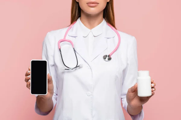 Vista recortada del médico sosteniendo teléfono inteligente con pantalla en blanco y frasco con pastillas aisladas en rosa - foto de stock