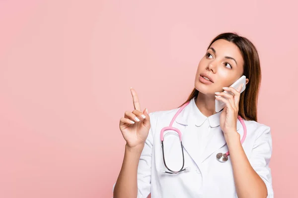 Médico con estetoscopio apuntando con el dedo mientras habla en smartphone aislado en rosa - foto de stock