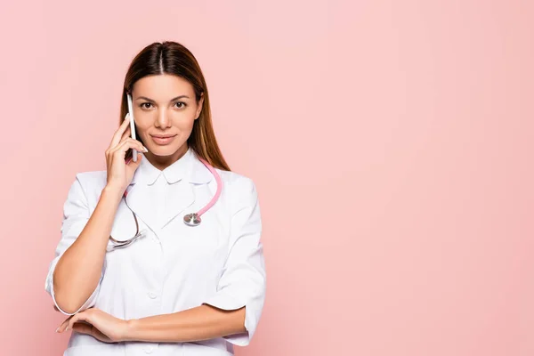 Médico sonriente hablando en el teléfono inteligente mientras mira la cámara aislada en rosa - foto de stock