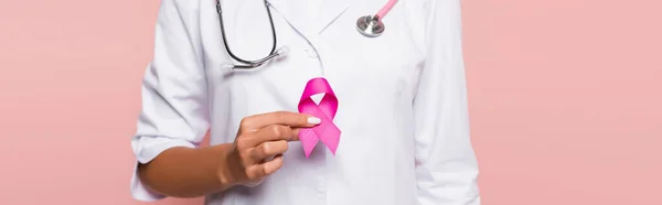 Vista recortada del médico en bata blanca que sostiene la cinta de la conciencia del cáncer de mama aislado en rosa, bandera - foto de stock